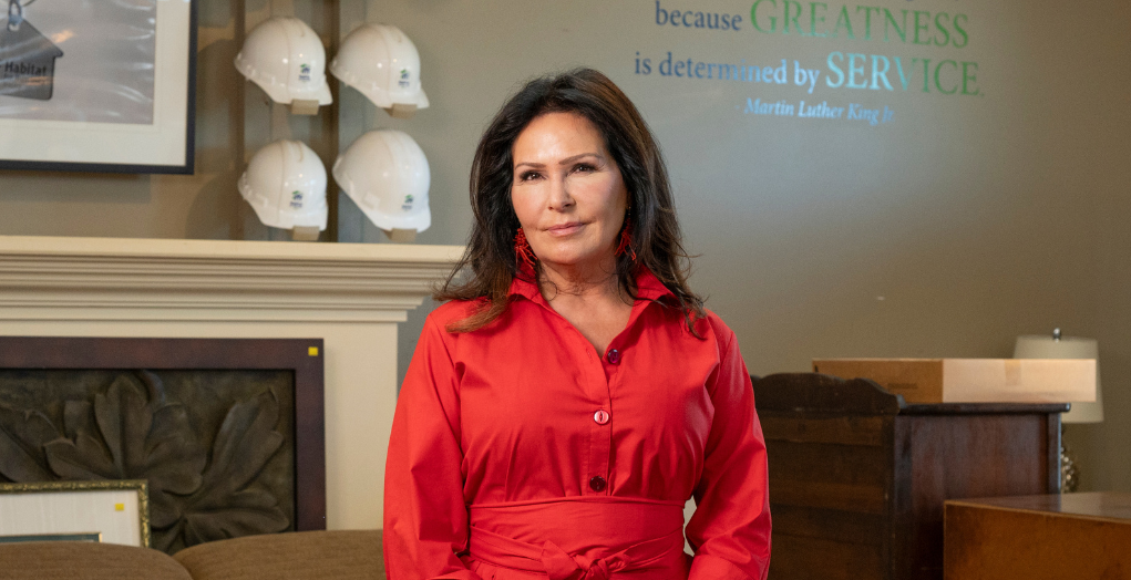 Cindi stands in the Fonthill ReStore in front of a wall with Habitat hardhats and a quote that says - because greatness is determined by service, Martin Luther King Jr. -- Cindi se tient dans le Fonthill ReStore devant un mur avec des casques Habitat et une citation qui dit - parce que la grandeur est déterminée par le service, Martin Luther King Jr.